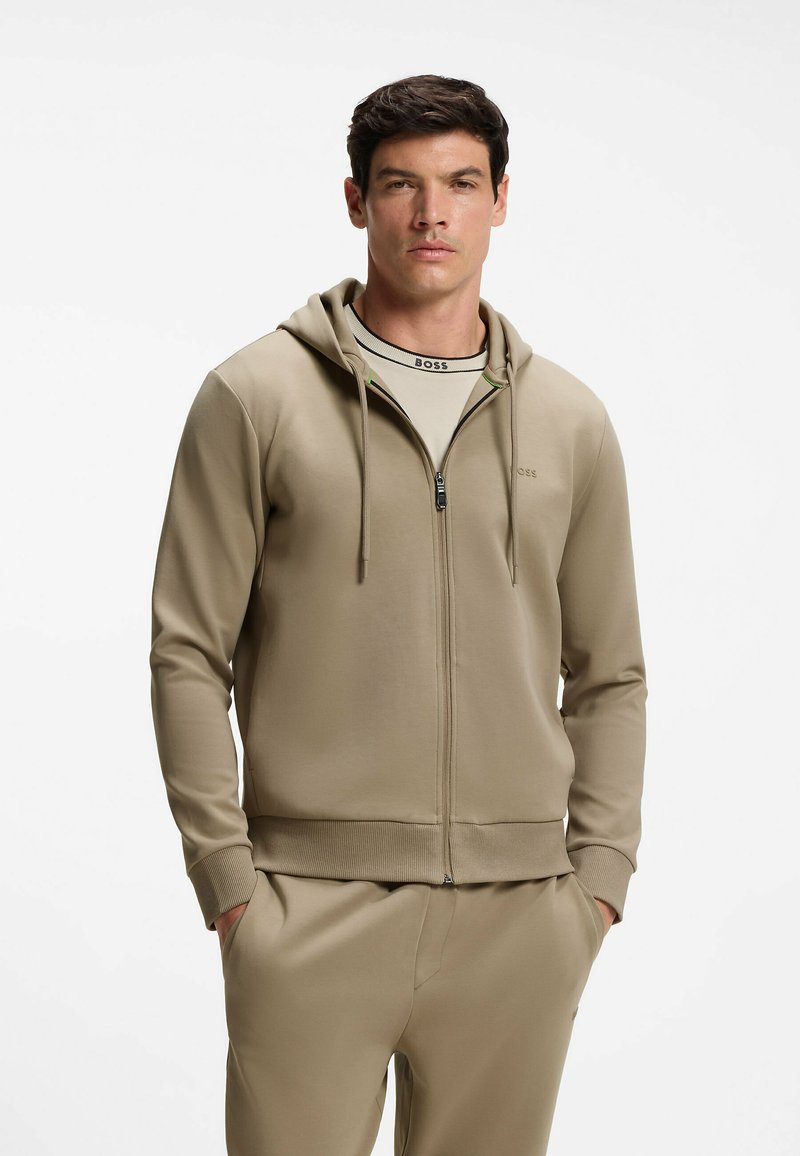 Homme portant un sweat à capuche beige zippé et un pantalon de survêtement assorti, les mains dans les poches, debout devant un fond blanc.