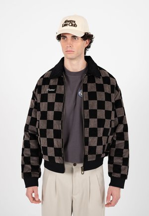 Muž nosící béžovou kšiltovku s nápisem "Always Late Club", černohnědou károvanou bundu na zip, šedé tričko a béžové plisované kalhoty stojící na bílém pozadí.