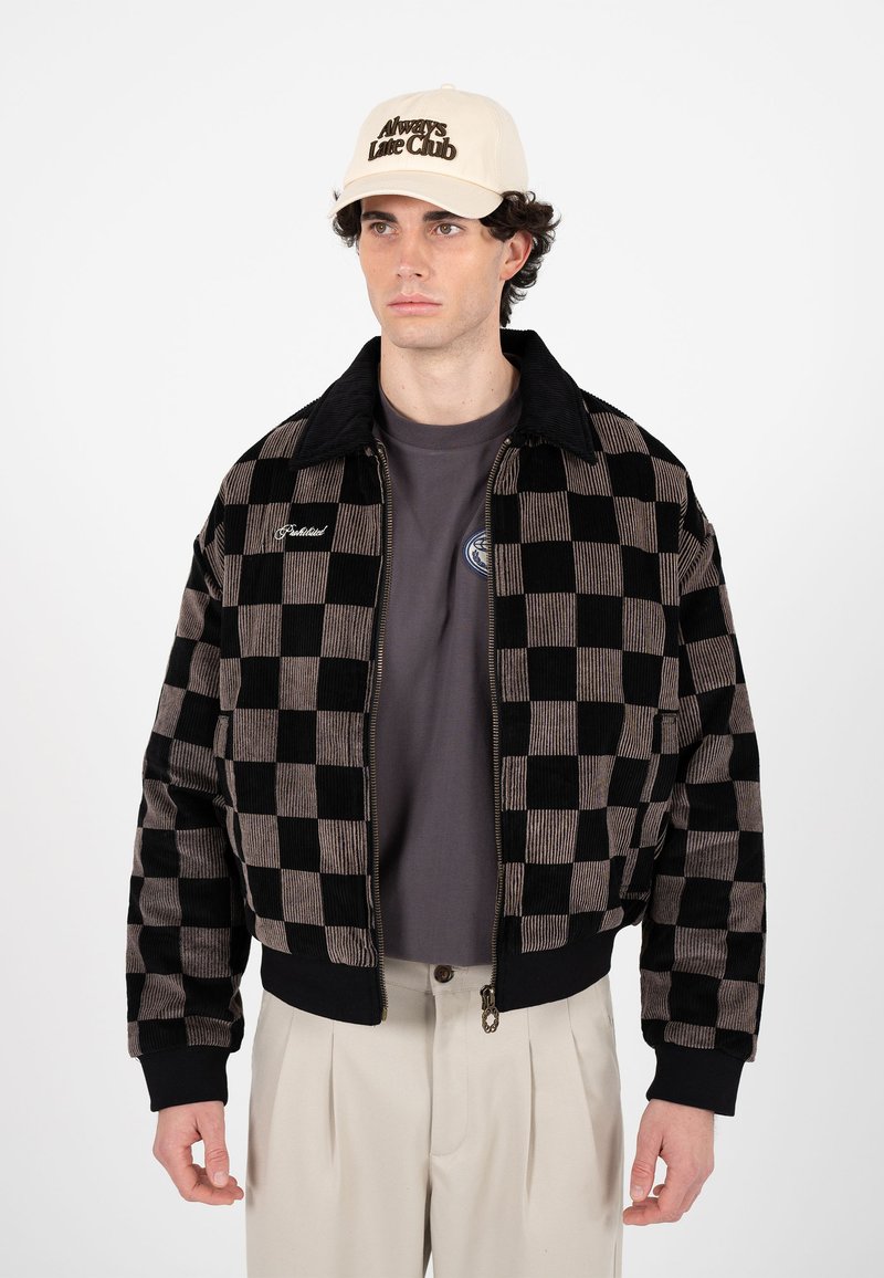 Homme portant une casquette beige "Always Late Club", une veste zippée à carreaux noirs et marron, un t-shirt gris et un pantalon plissé beige, debout devant un fond blanc.