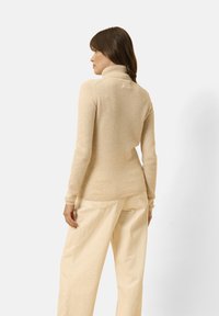 Maglione a collo alto beige realizzato in tessuto morbido e testurizzato. Presenta maniche aderenti e un orlo dritto, abbinato a pantaloni larghi di colore chiaro.