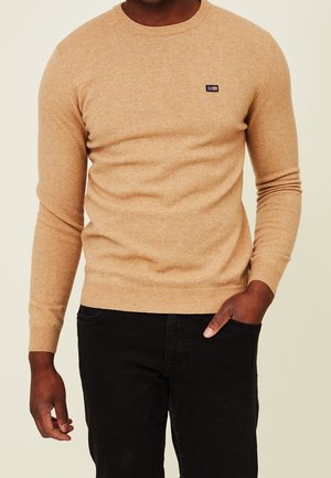 Mand iført en beige crewneck-sweater med et lille sort logo på brystet og sorte bukser, den ene hånd i lommen.