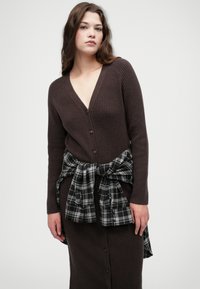 Καφέ ριγέ καρντιγκάν με βαθύ V-λαιμόκοψη, κουμπωτή μπροστινή μεριά και μακριά μανίκια, συνδυασμένο με πουκάμισο σε ασπρόμαυρη καρό παραλλαγή που είναι δεμένο γύρω από τη μέση.