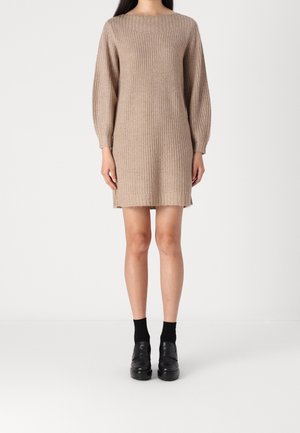 Vrouw draagt een lange mouw, knie-lange beige gebreide jurk met zwarte sokken en zwarte platformschoenen, staand tegen een effen achtergrond.