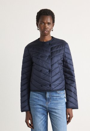 Marineblå quiltet jakke med rund hals, dobbeltbrettede knapper og chevronbroderi, paret med lyseblå jeans.