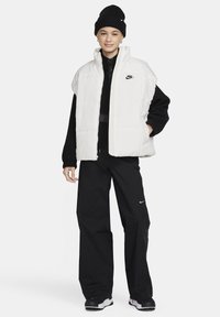 Biała pikowana kamizelka z wysokim kołnierzem i zamkiem błyskawicznym. Nosi się ją na czarny sweter o fakturze i czarne spodnie. Całość dopełniają czarna czapka typu beanie i czarne sneakersy.