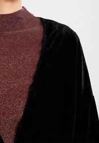 Personne portant un col roulé en maroon pailleté sous une veste en velours noir, vue du cou à mi-torse sur un fond uni.