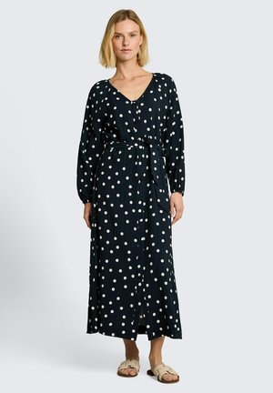 Femme portant une longue robe noire à pois blancs avec une ceinture à la taille, debout et chaussée de sandales beiges sans bride, sur un fond uni.