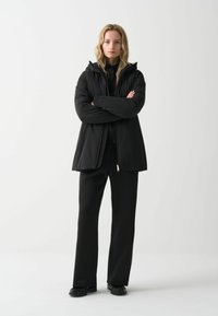 Zwarte gewatteerde jas met capuchon, ritssluiting en lange mouwen, gecombineerd met zwarte wijde broek en platte schoenen. Glad, mat textuur.