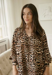 Túnica con estampado de leopardo en tela suave, con escote en V con lazos, mangas largas y corte holgado. Los colores incluyen marrón, negro y crema.