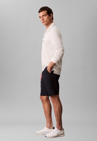 Tricou polo alb cu mânecă lungă și trei nasturi, asortat cu pantaloni scurți de culoare albastru închis și adidași albi, stând pe un fundal gri.