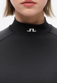 Κοντινό πλάνο ενός ατόμου που φοράει μαύρο αθλητικό μπλουζάκι με ψηλό γιακά και ένα μικρό λευκό γεωμετρικό λογότυπο στο κέντρο του γιακά.