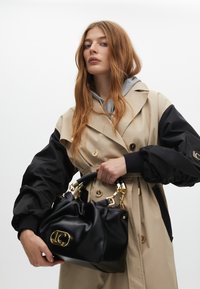 Czarna torebka z miękką, marszczoną strukturą i złotymi akcentami, trzymana przez osobę w beżowym i czarnym płaszczu z dużymi guzikami.