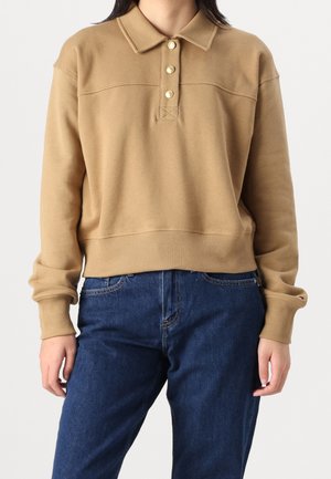 Osoba nosząca camelowy sweter typu polo z długim rękawem z trzema ozdobnymi guzikami oraz niebieskie dżinsy, stojąca na jednolitym tle.