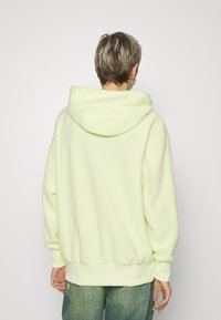 Sudadera de felpa amarilla clara con una gran capucha, corte relajado y puños acanalados. Combinada con jeans azules de ajuste holgado. Detalles de diseño mínimos visibles.