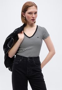 Νεαρή γυναίκα που φοράει μαύρο και λευκό ριγέ μπλουζάκι με V λαιμόκοψη και μαύρο τζιν ψηλόμεσο, κρατώντας ένα μαύρο σακάκι στον ώμο της, πάνω σε λευκό φόντο.