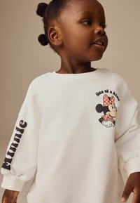 Vit sweatshirt med långa ärmar som har en lekfull grafisk figur på framsidan och text längs ärmen i feta svarta bokstäver.