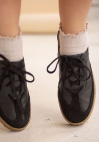 Mustat nahka- ja mokkakengät pyöreällä kärjellä ja nauhallisella mallilla, yhdistettynä valkoisiin resorillisiin nilkkasukkiin, joissa on aaltoileva reuna.