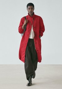 Roter langer Mantel mit hohem Kragen, Knopfverschluss und Seitentaschen, getragen über einem hellen Oberteil und schwarzen weiten Hosen. Dazu Stiefeletten.