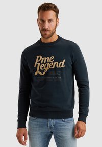 Mornarica pamučni dukserica s zlatnim otisnutim tekstom "Pme Legend" i manjim tekstom ispod. Ima rebraste manžete i rub, kao i okrugli izrez.