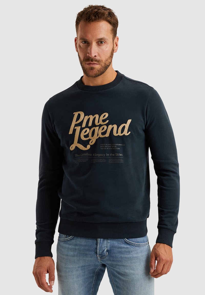 Mornarica pamučni dukserica s zlatnim otisnutim tekstom "Pme Legend" i manjim tekstom ispod. Ima rebraste manžete i rub, kao i okrugli izrez.