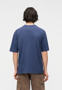 Άτομο με κοντά σκούρα μαλλιά που φοράει απλό μπλε κοντομάνικο μπλουζάκι και καφέ παντελόνι cargo, απεικονίζεται από την πίσω πλευρά σε ανοιχτόφωρο φόντο.