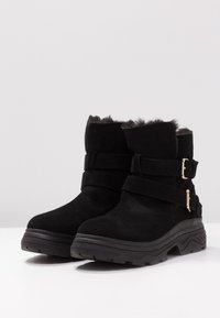 Botas negras de tobillo de ante con forro de piel de pelo, que presentan dos correas anchas y hebillas metálicas. Suela de goma resistente con un diseño robusto.