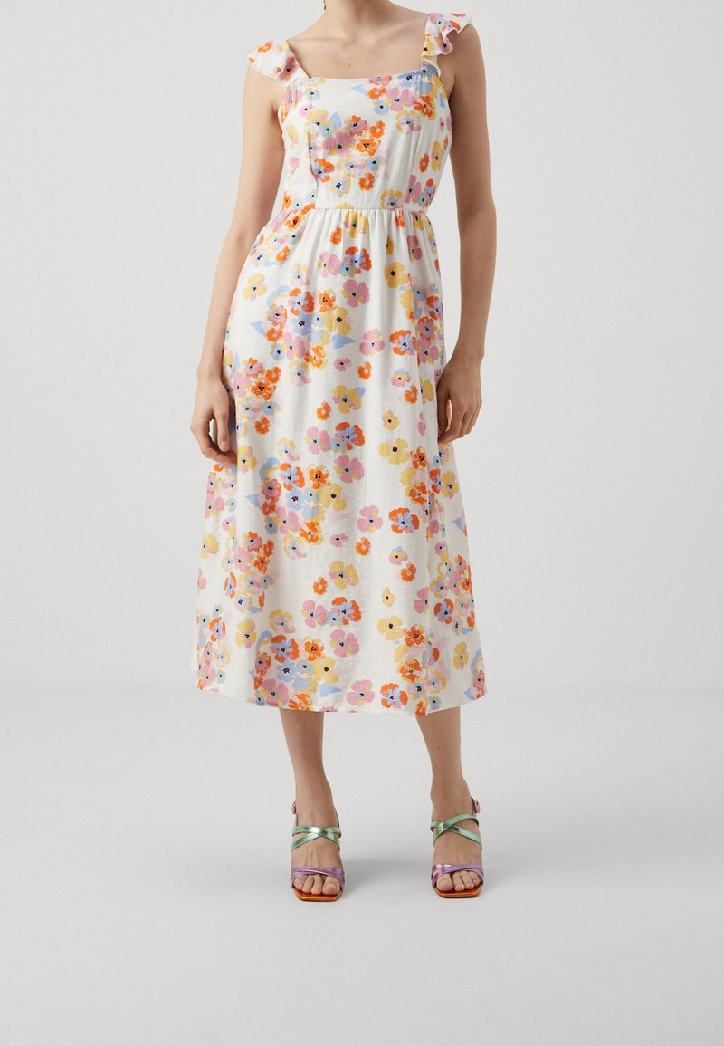 Femme portant une robe midi blanche avec un imprimé floral coloré et des bretelles volantées, associée à des sandales multicolores à brides.