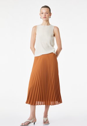 Femme portant un haut blanc côtelé sans manches, une jupe midi plissée couleur rouille, des boucles d'oreilles argentées en forme d'anneau et des sandales blanches à talons bas ouvertes.