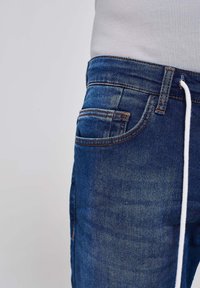 Jeans iz temno modrega jeansa s subtilnim zbledeljem, ki imajo pet žepov, okrasne šive in pas z vrvico.