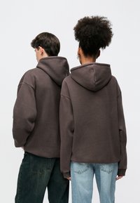 Felpe marroni con vestibilità rilassata, caratterizzate da cappucci oversize e polsini a coste, indossate da due persone, abbinate a jeans blu e verde scuro.