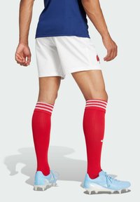 Shorts blancs, haut de sport bleu, chaussettes rouge jusqu'au genou avec des rayures blanches, et chaussures de soccer bleu clair avec un design texturé.