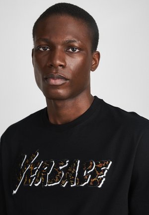 Fekete pulóver, amelyen a "VERSACE" logó texturált, színes hímzéssel, enyhe fényességgel látható. Kerek nyakú dizájn, minimalista esztétika.