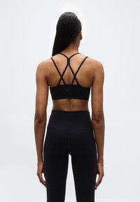 Γυναίκα που φαίνεται από πίσω φοράει μαύρο αθλητικό σουτιέν με ιμάντες και μαύρο ψηλόμεσο κολάν, σε απλό φωτεινό φόντο.