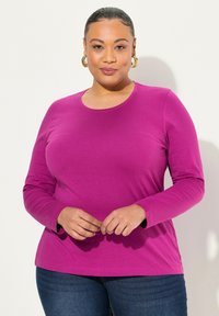 Langarmshirt mit Rundhalsausschnitt aus lebhaftem Fuchsia-Stoff. Glatte Textur mit körpernaher Passform, kombiniert mit dunkelblauen Jeans.