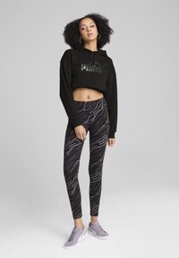 Zwarte cropped hoodie met een reflecterend logo, gecombineerd met zwarte leggings met een grijs golvend patroon. Schoeisel zijn lichtpaarse sneakers.