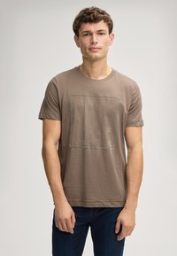 Bruine katoenen t-shirt met korte mouwen, voorzien van een geometrisch patroon aan de voorkant. Ronde halslijn en een losse pasvorm. Gdraagd met blauwe jeans.