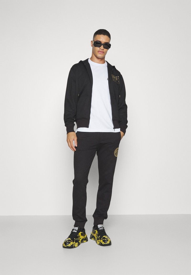 Zwarte zip-up hoodie met gouden accenten, wit t-shirt, zwarte joggers met een logo, en zwarte sneakers met gele patronen.