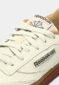 Plan rapproché d'une basket blanche Reebok Classic avec doublure intérieure beige, lacets blancs et semelle marron, arborant le logo Reebok et un patch du drapeau britannique.