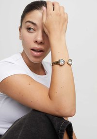 Pulsera de oro con dos gemas azules triangulares, textura suave, llevada en la muñeca. Persona con camisa blanca, posando con expresión neutra.