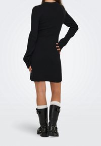 Femme portant une robe noire à manches longues côtelée, des chaussettes blanches et des bottes noires à boucles, debout de dos face vers l'avant.