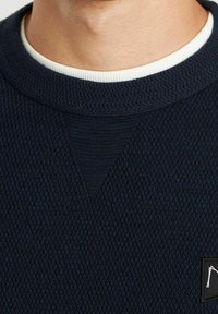 Pull texturé bleu marine avec un col rond, un col accent blanc et une petite étiquette logo sur le côté gauche.