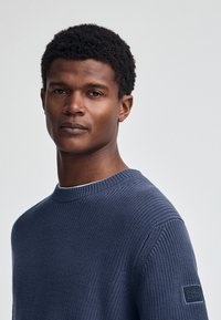 Marineblauwe gebreide trui met een ronde hals, voorzien van een textuurpatroon en een klein zwart "POLO CLUB"-label op de linkermouw.