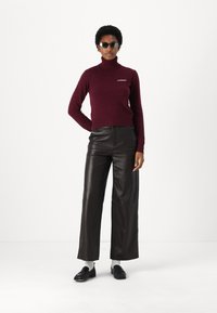 Bordo turtleneck megztinis su siuvinėtu logotipu, derinamas su juodomis odinėmis plačiomis kelnių iki juosmens, ir juodais loaferiais.
