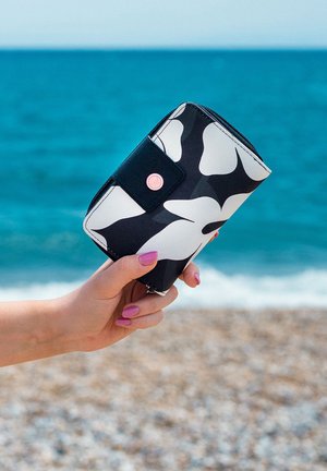 Black and white patterned wallet with a zipper and snap closure, featuring a pink accent button, held against a beach background.
