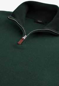Maglione verde scuro con zip al collo realizzato in tessuto a maglia testurizzata. Presenta una zip metallica con un tirante in pelle marrone e un colletto a costine.