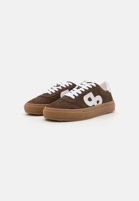 Baskets en suède marron avec des lacets blancs et un logo blanc surdimensionné. Présentent une semelle en caoutchouc texturée et un col rembourré en beige clair.