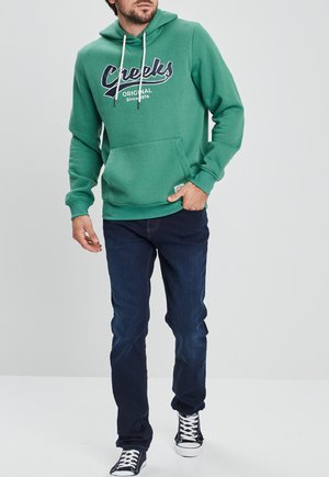Groene hoodie met witte trekkoorden en een voorzak, met marineblauwe letters. Draagt het met donkerblauwe jeans en zwart-witte hoge sneakers.