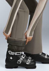 Scarponi da sci neri con fibbie argentate. Pantaloni oliva con strisce laterali beige chiare e polsini elastici, aderenti agli scarponi.