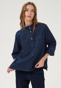 Tamsiai mėlynas džinsinis marškinis su henley apykakle, auksinėmis mygtukais, dviem krūtinės kišenėmis ir trijų ketvirčių rankovėmis. Minkšta tekstūra ir kasdieniškas dizainas.