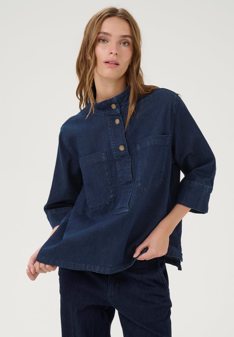 Navyblauwe denim overhemd met een henley-kraag, voorzien van gouden knopen, twee borstzakken en driekwartmouwen. Zachte textuur en casual ontwerp.